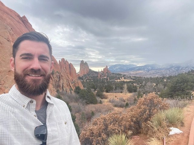 A bearded man wearing a white checkered shirt standing in front of a scenic rocky landscape with mountains and trees in the background.