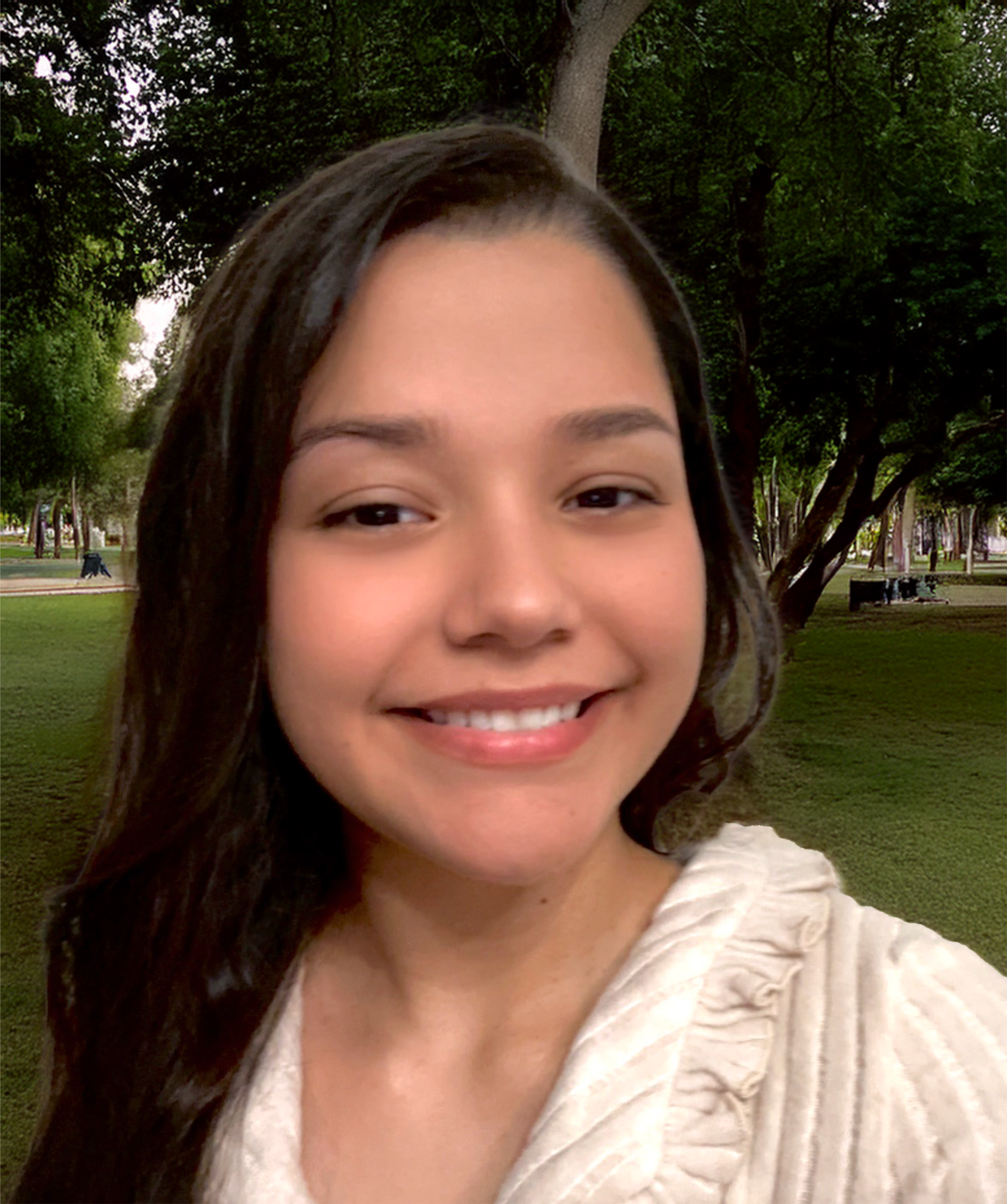 A smiling woman with long dark hair, wearing a light-colored blouse with ruffles, standing in a park with green trees in the background.
