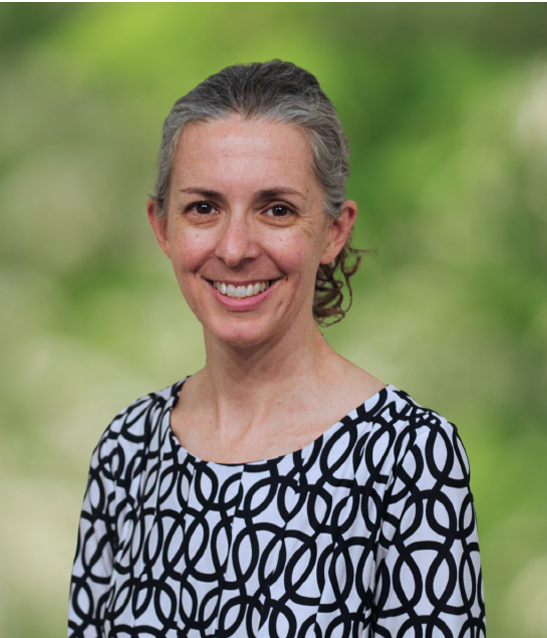 A woman with gray hair pulled back, wearing a patterned black and white blouse, smiling against a blurred green background.