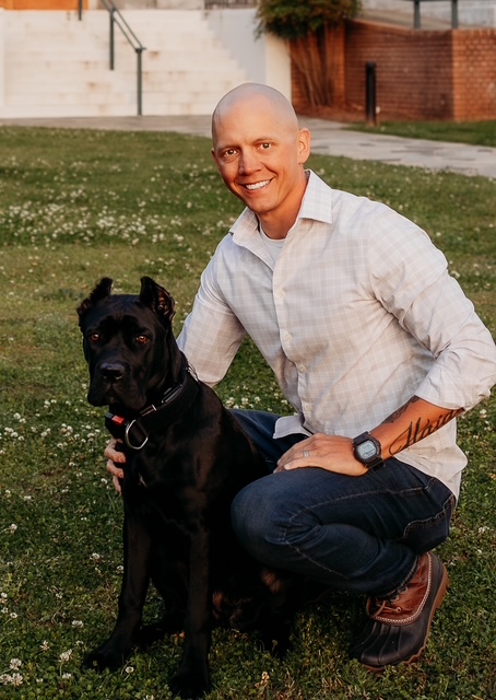 A bald man in a white shirt kneeling on the grass with a black Cane Corso dog, smiling at the camera.