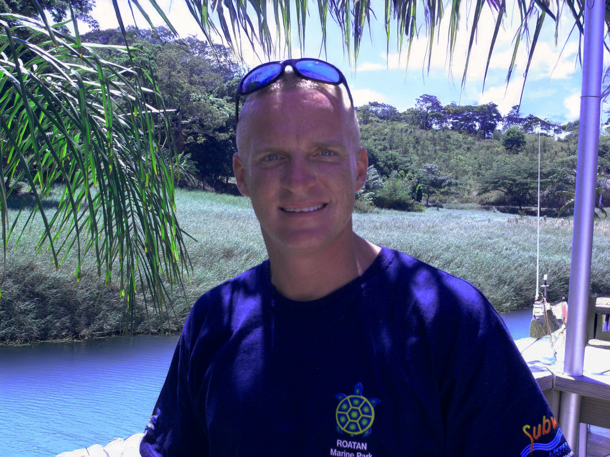 A bald man wearing sunglasses on his head and a navy blue shirt with a marine logo, standing outdoors near a body of water with palm leaves and greenery in the background.