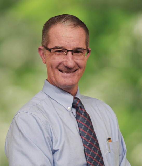 A professional-looking man wearing glasses, a blue shirt, and a tie, smiling against a blurred green background.